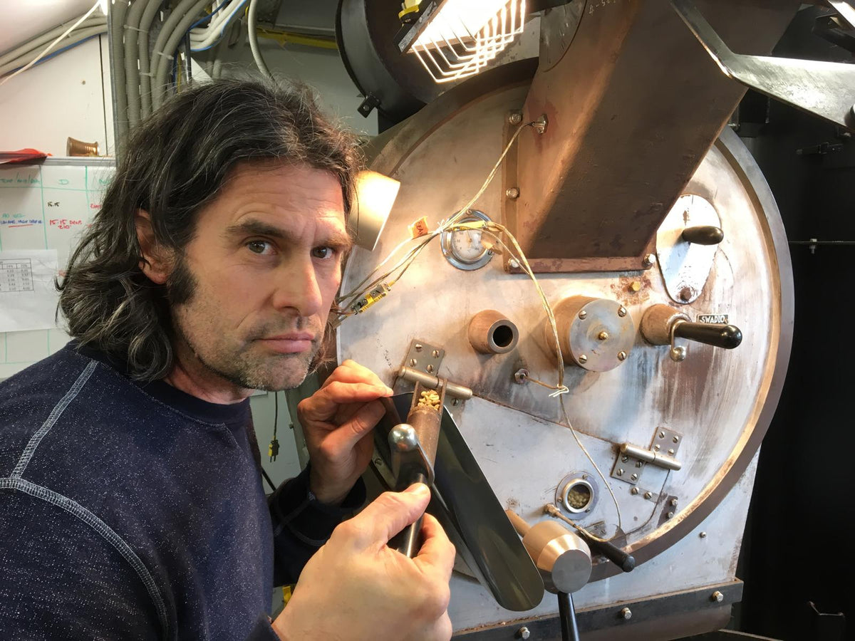 A man with long dark hair (Jason) works on a coffee roaster machine inside what looks like a workshop or laboratory. The machine has copper-colored upper sections and silver metal components, with wires and small hardware visible.