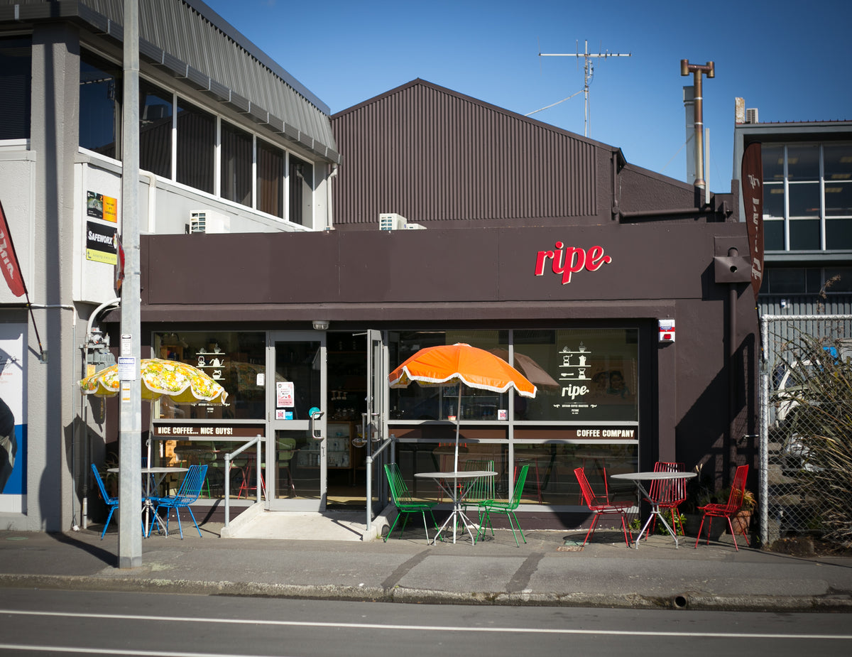A street-facing view of a dark brown storefront with a red 'ripe' sign, outdoor seating with colorful chairs and umbrellas, and a clear blue sky above.