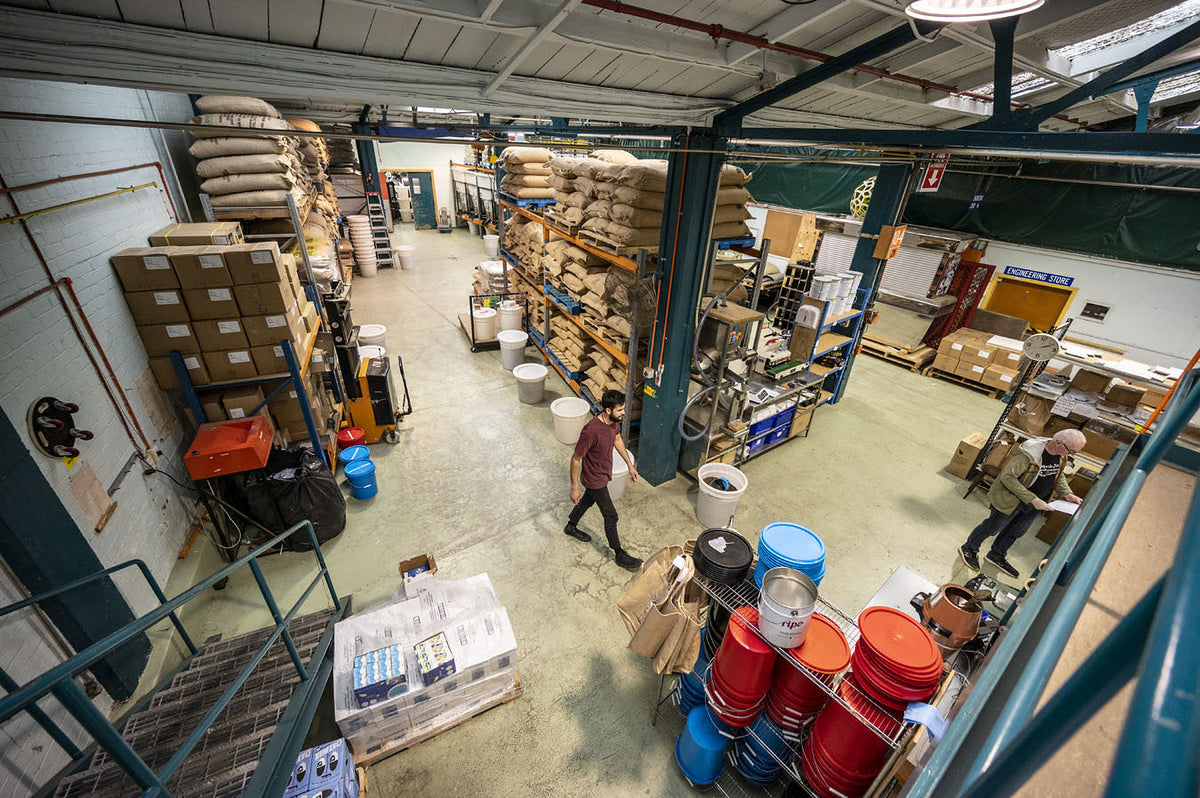 An overhead view of a large warehouse interior with shelving, stacked boxes, bags, buckets, and two people walking through the space.